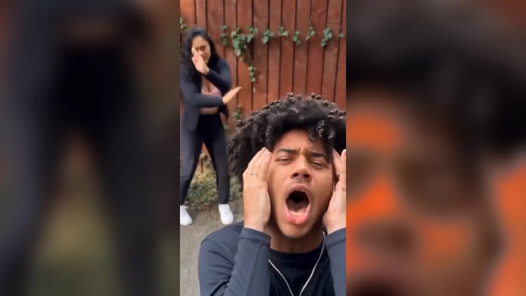 A young man with curly hair screams with both hands on his head in front of a wooden fence while a woman dances behind him